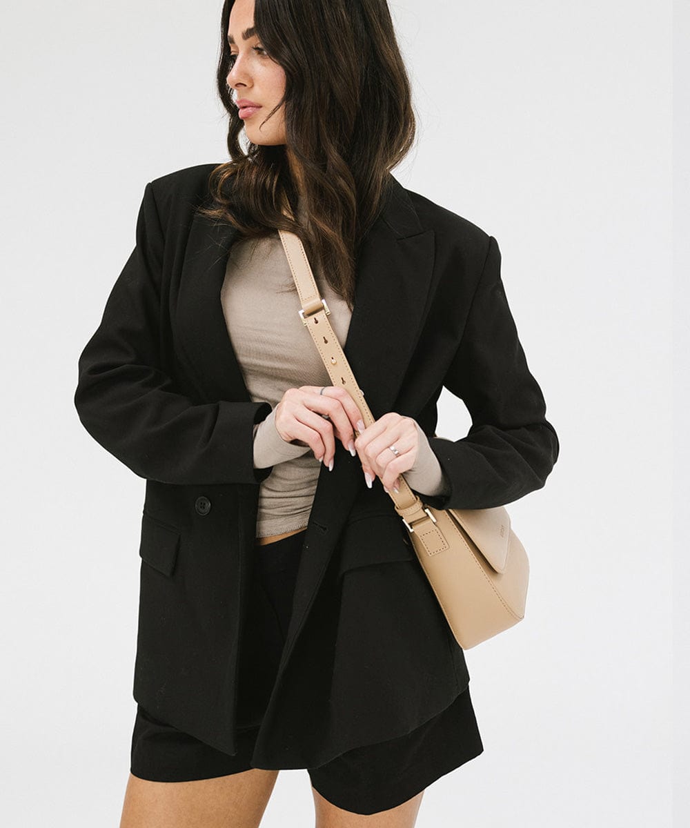 Woman wearing a black blazer and black shorts, carrying a tan Rhys Crossbody Genuine Leather Bag against a plain background #color_tan