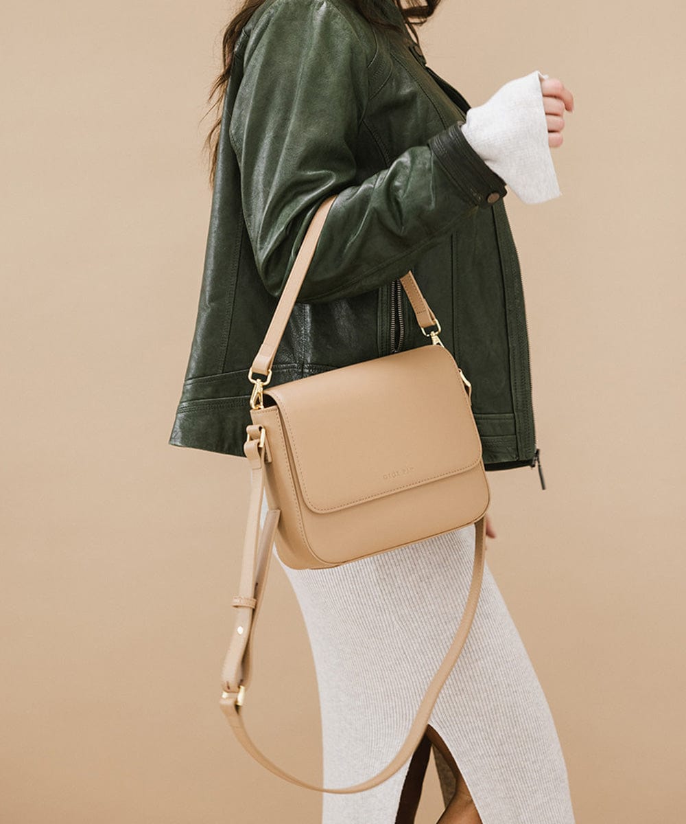 Woman wearing a dark green jacket and a white skirt, carrying a tan Rhys Crossbody Genuine Leather Bag, posing sideways against a plain background #color_tan
