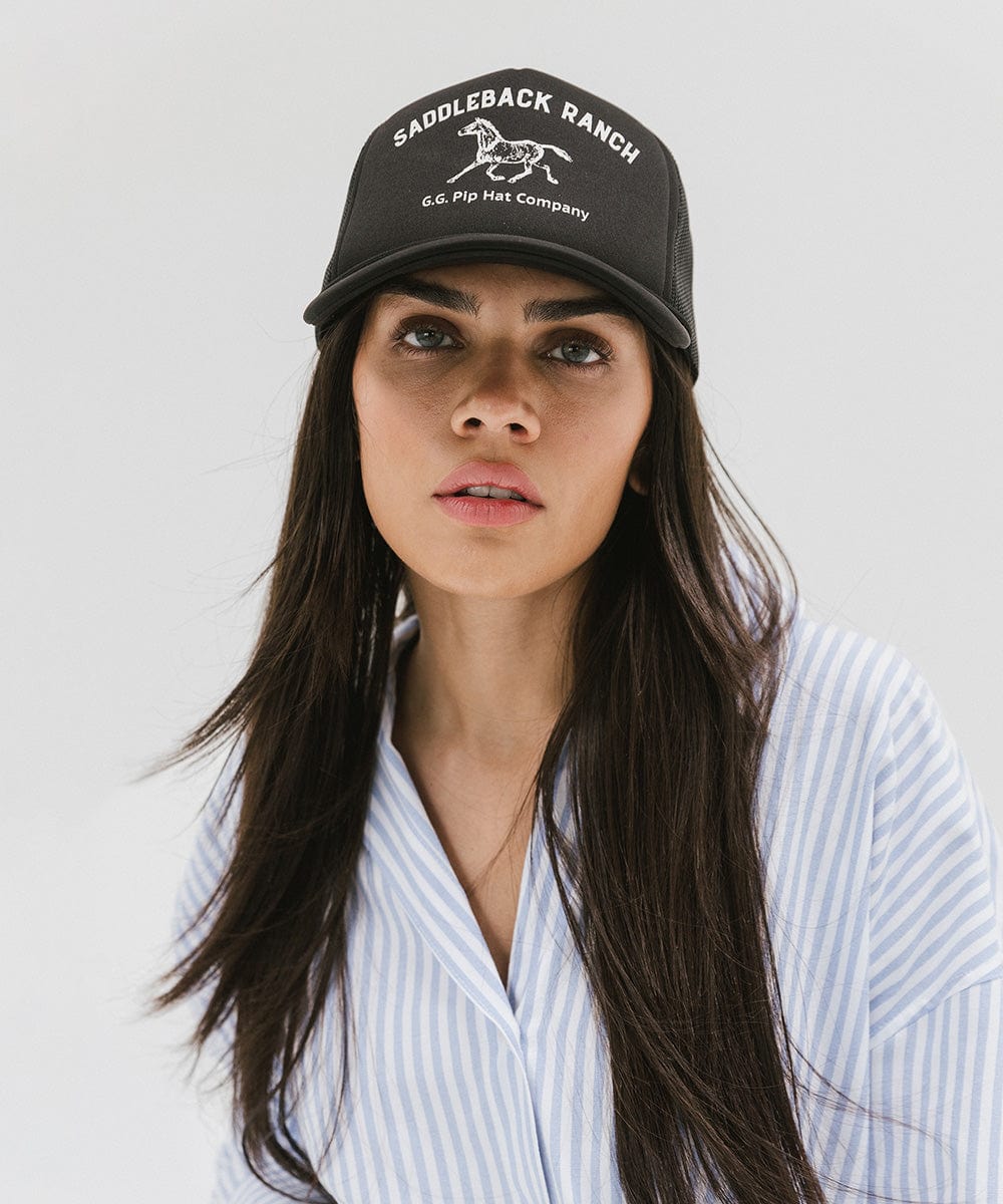 Woman wearing a black cap with 'Saddleback Ranch' text and logo and a light colored shirt against a plain background #color_black