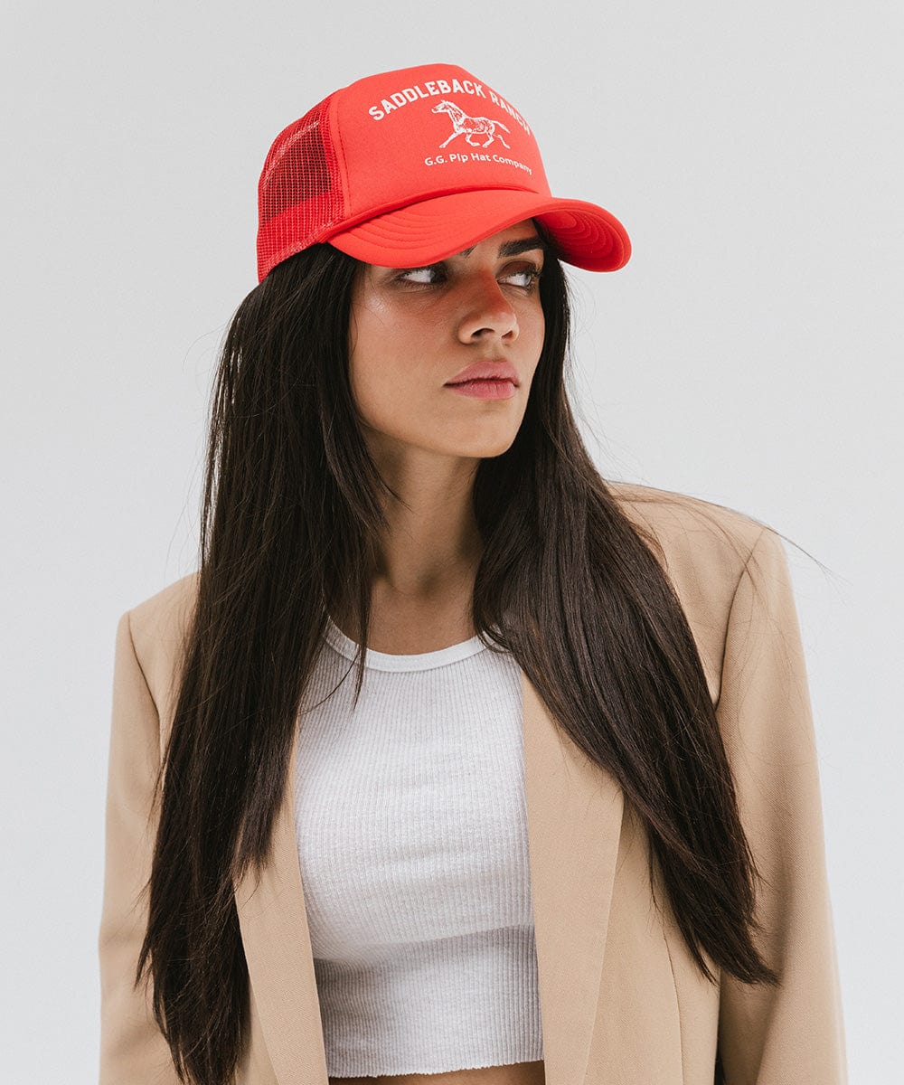 Woman wearing a vintage red colored cap with 'Saddleback Ranch' text and logo, a white top and beige coat against a plain background #color_vintage red
