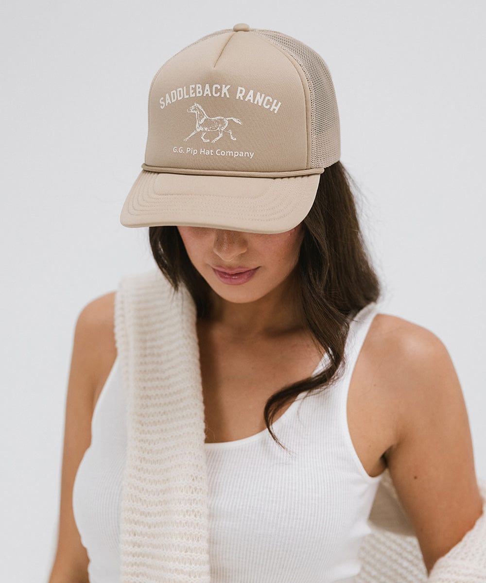 Woman wearing a tan cap with 'Saddleback Ranch' text and logo, and a white top, posing against a plain background #color_tan