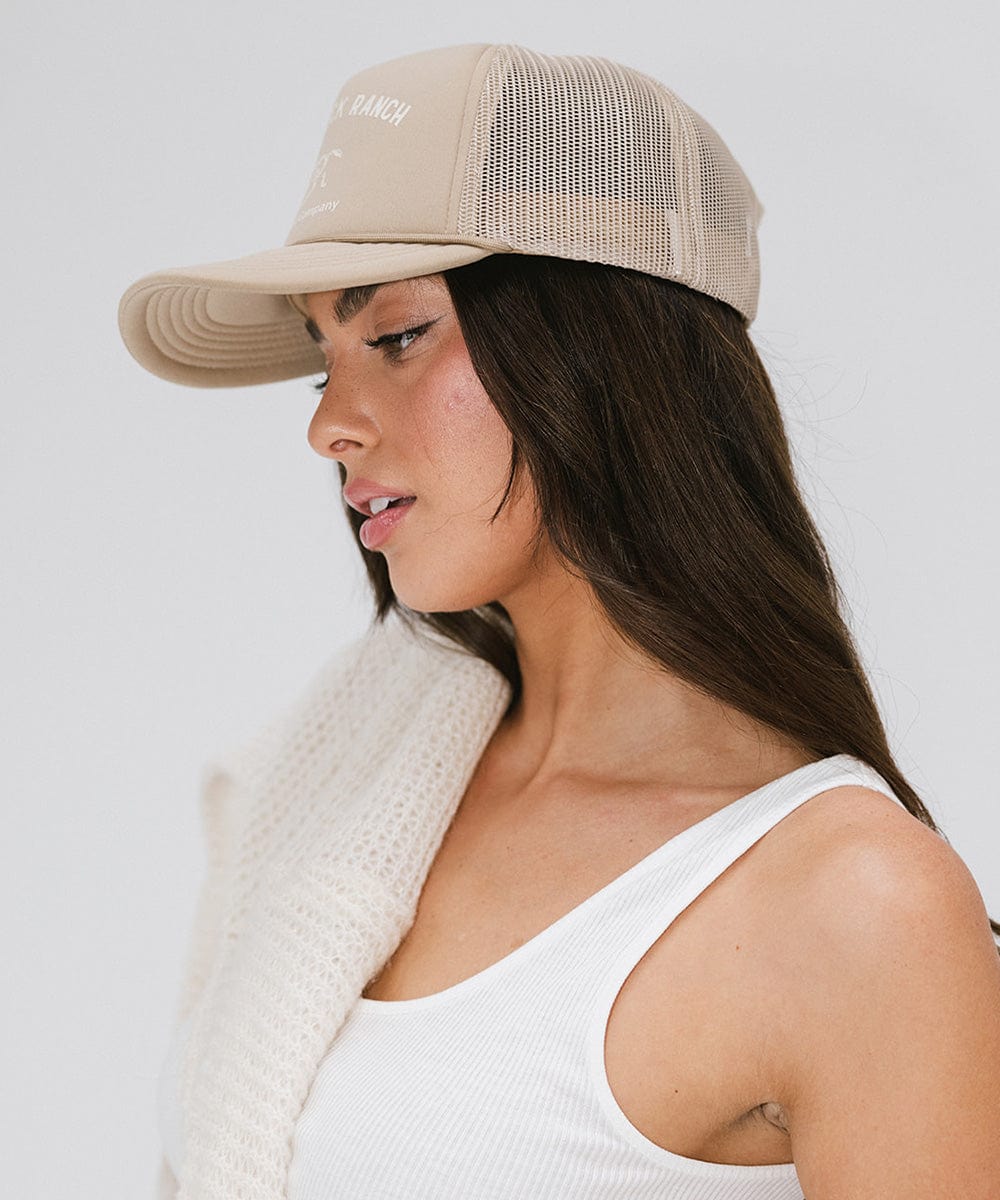 Woman wearing a tan cap with 'Saddleback Ranch' text and logo, and a white top, posing sideways against a plain background #color_tan