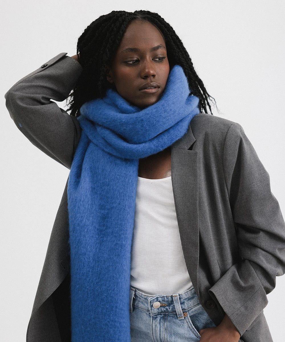 A woman wearing a grey blazer and an alpine blue colored oversized knit scarf with fringes, draped over the neck against a plain background #color_alpine blue