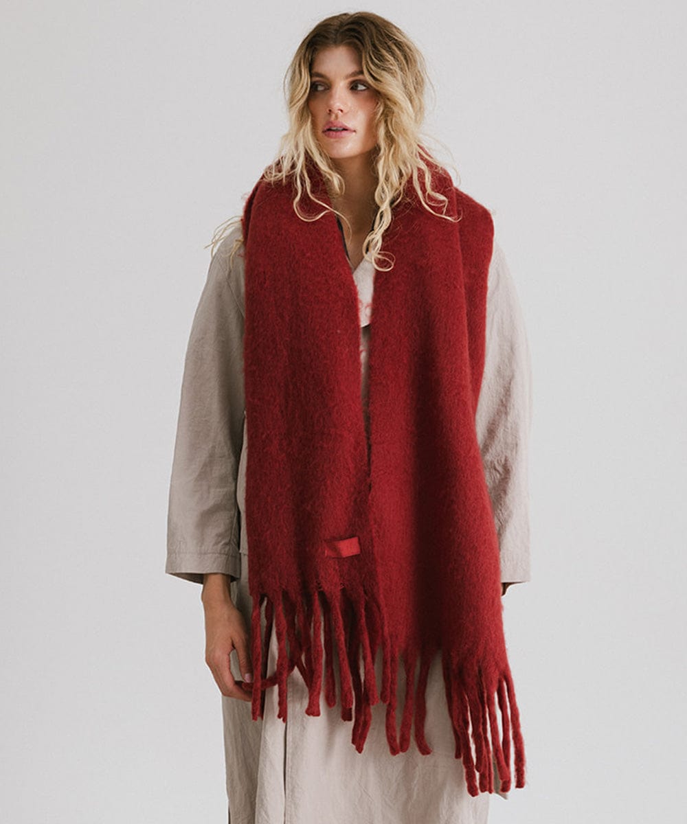 Woman wearing a burgundy oversized knit scarf with fringes, draped over the neck against a plain background #color_burgundy