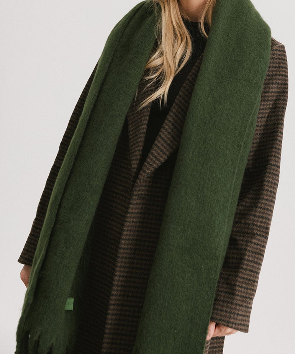 A person wearing a coat and a dark green oversized knit scarf with fringes, draped over the neck against a plain background #color_dark green