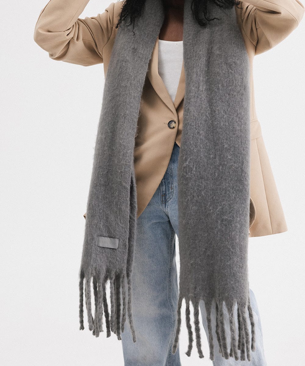 A person wearing a beige blazer and blue jeans, and a grey oversized knit scarf with fringes, draped over the neck against a plain background #color_grey