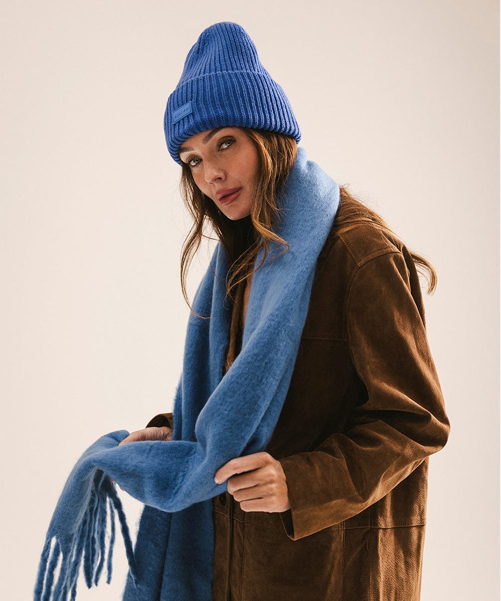 Woman wearing a blue knit hat and scarf against a plain background #color_alpine blue