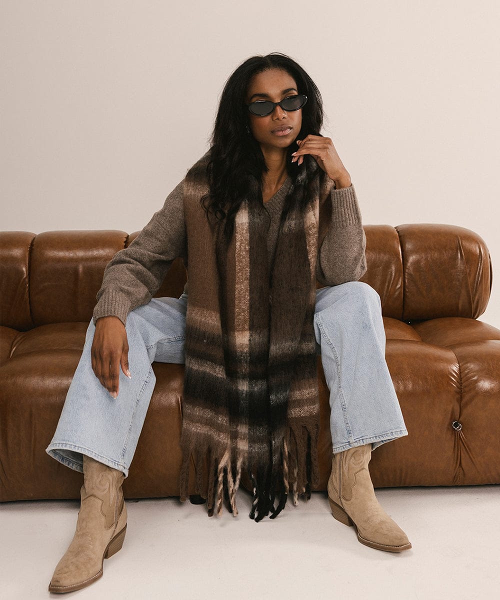 Woman sitting on a brown leather couch wearing sunglasses, a brown sweater, light blue jeans, and beige boots. #color_Brown - Black Plaid