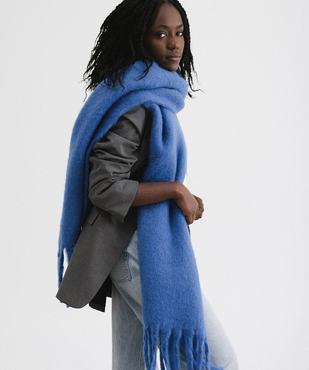 A woman wearing a grey blazer and blue jeans, and an alpine blue colored oversized knit scarf with fringes, draped over the neck against a plain background #color_alpine blue