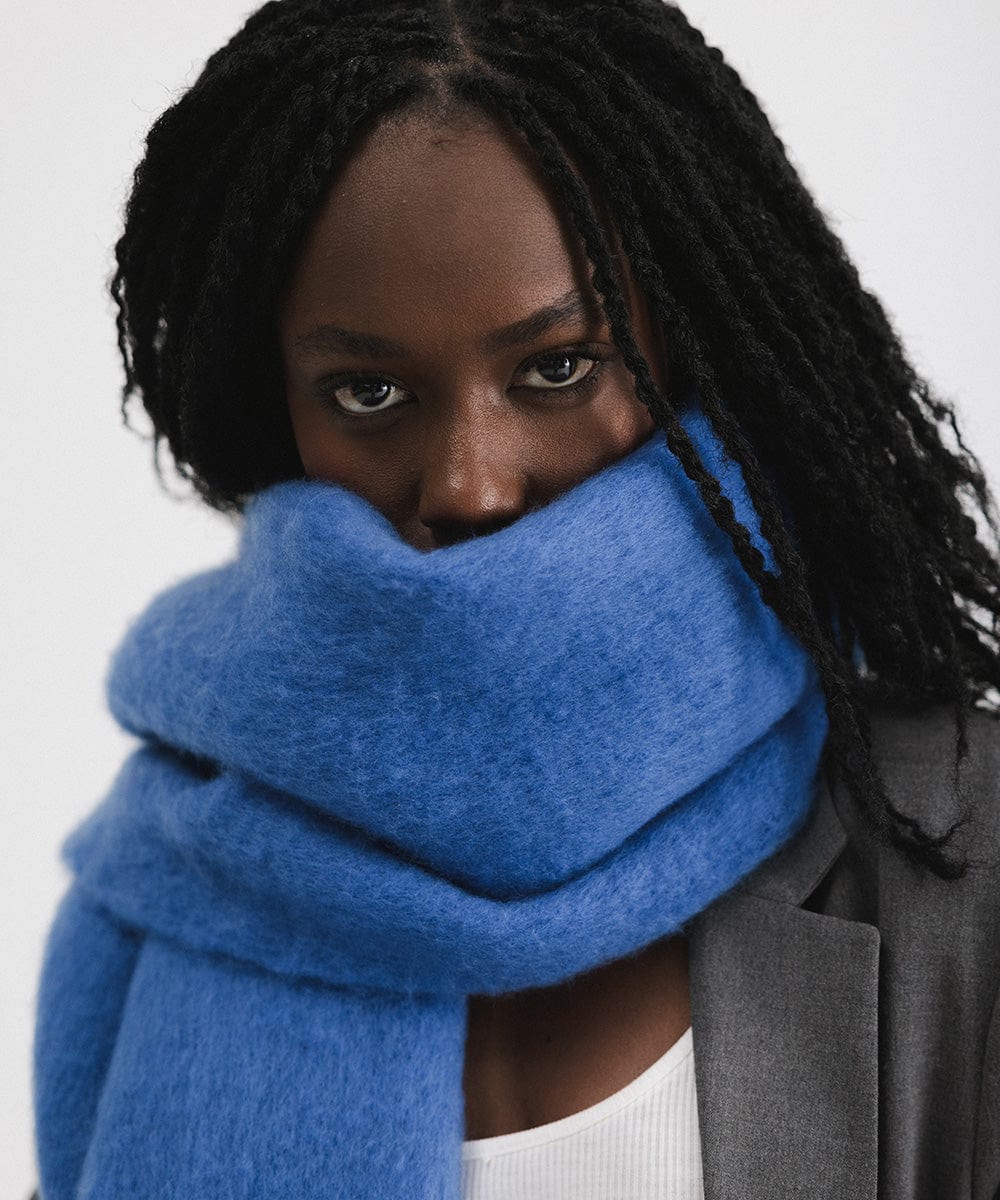 A person wearing an alpine blue colored oversized knit scarf with fringes, draped over the neck against a plain background #color_alpine blue