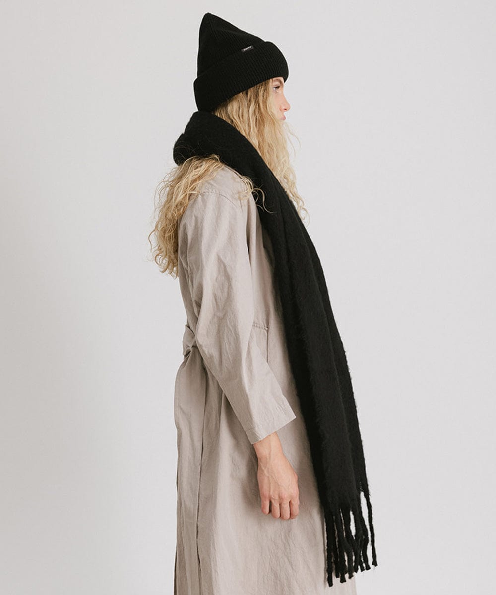 A person wearing a black beanie, a cream coat, and a black oversized knit scarf with fringes, draped over the neck against a plain background #color_black