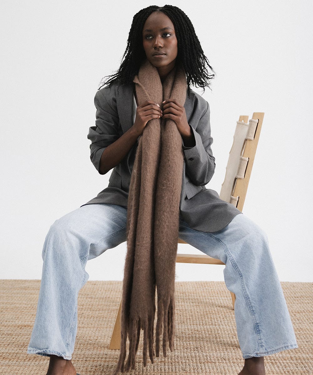A person wearing a grey blazer and blue jeans, and a brown oversized knit scarf with fringes, draped over the neck against a plain background #color_brown
