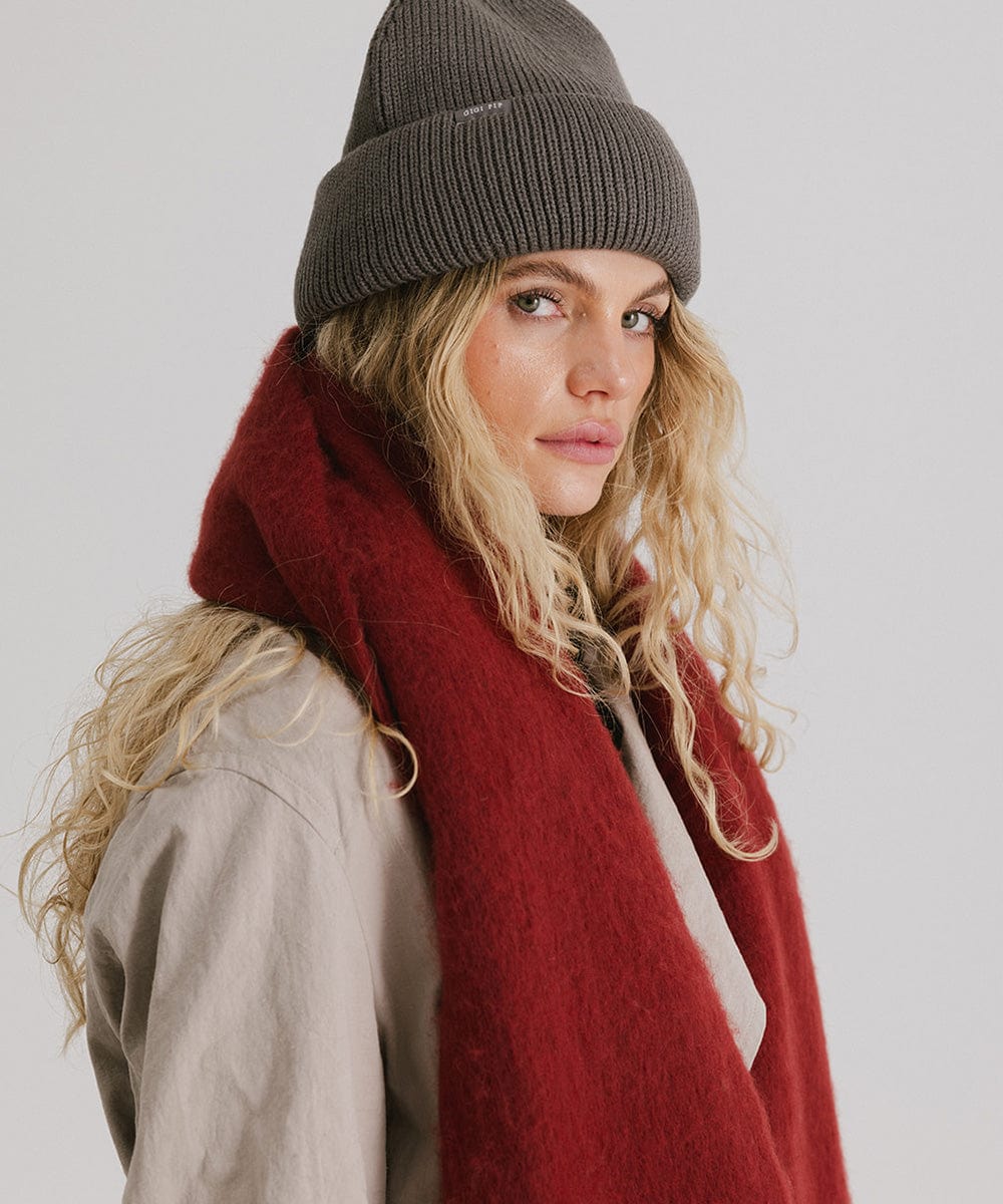 Woman wearing a grey beanie and a burgundy oversized knit scarf with fringes, draped over the neck against a plain background #color_burgundy