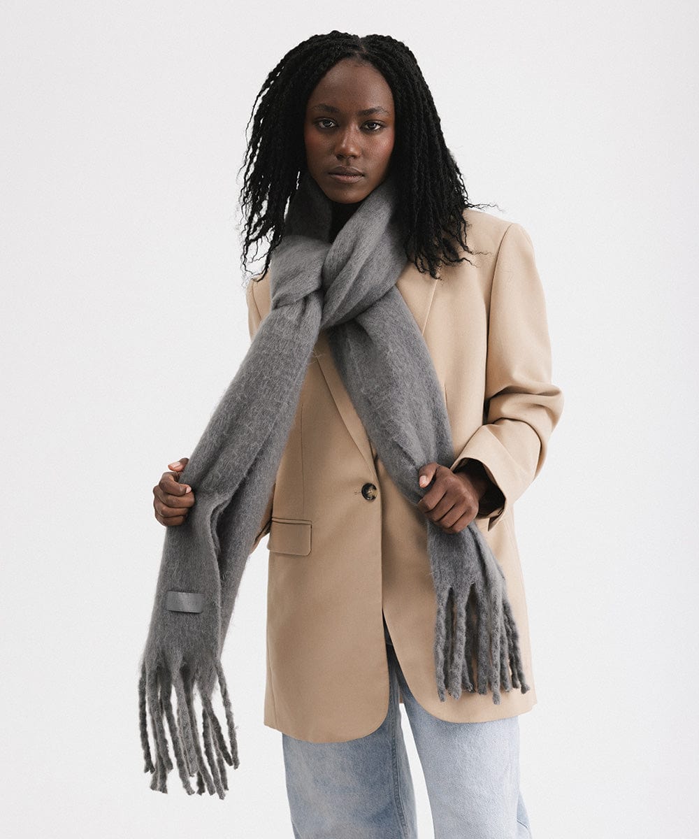 A woman wearing a beige blazer and blue jeans, and a grey oversized knit scarf with fringes, draped over the neck against a plain background #color_grey