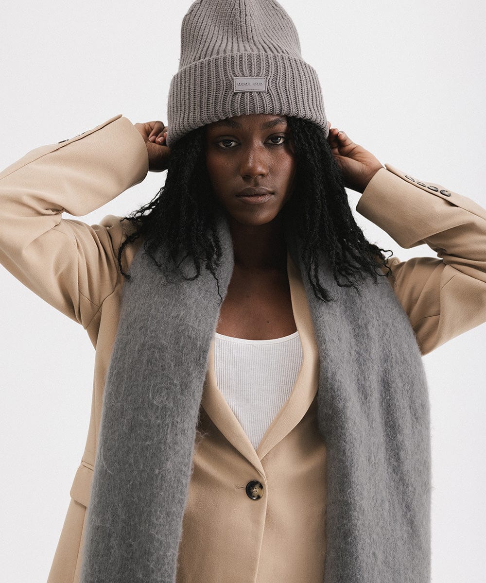 A person wearing a beige blazer and grey beanie, and a grey oversized knit scarf with fringes, draped over the neck against a plain background #color_grey
