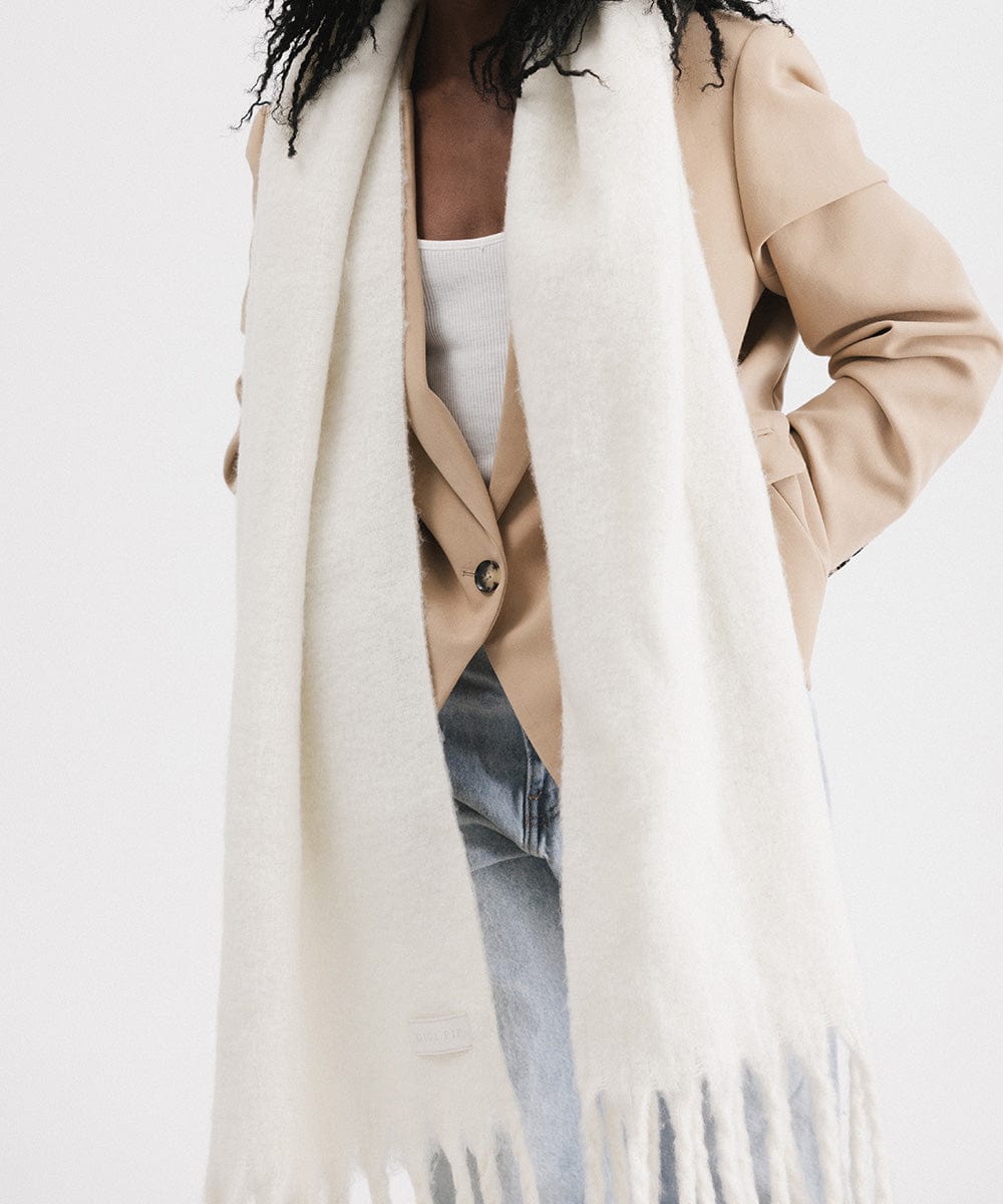 A person wearing a beige blazer, blue jeans, and an off white oversized knit scarf with fringes, draped over the neck against a plain background #color_off white