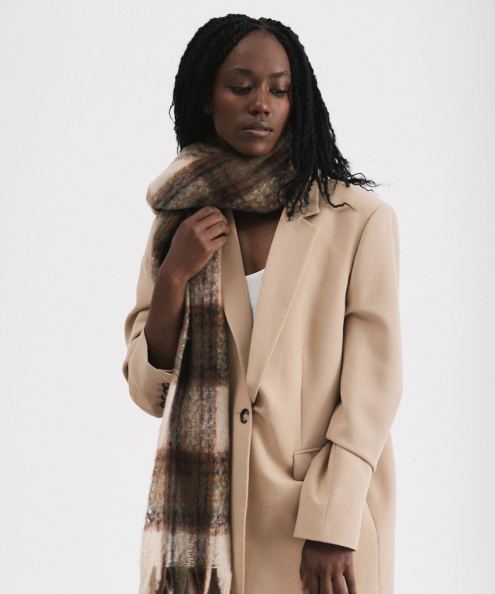 A woman wearing a coat, and a cream and tan colored oversized knit scarf with fringes, draped over the neck against a plain background #color_cream - tan