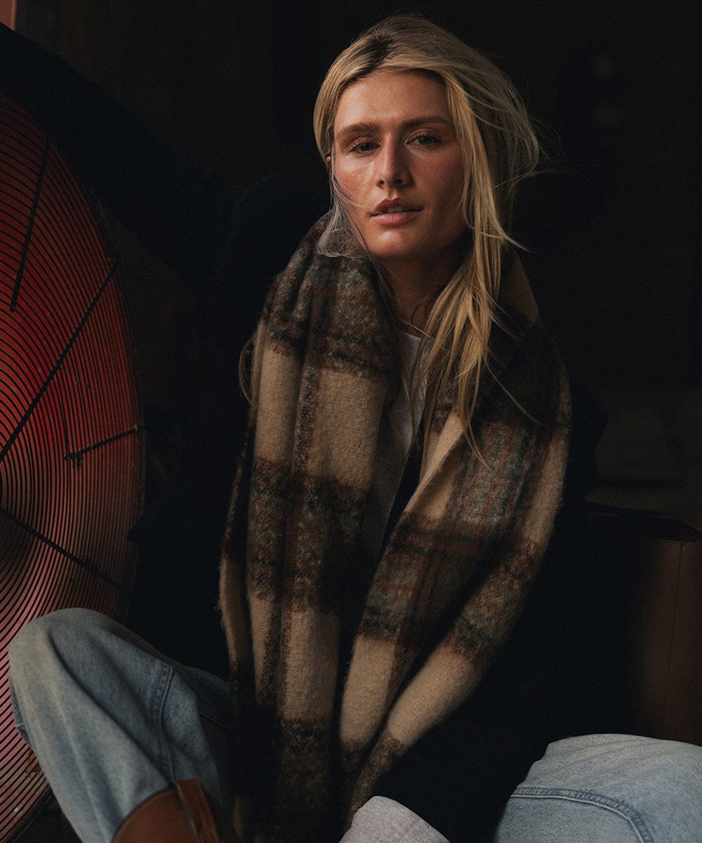 A person wearing a black coat and a cream and tan colored oversized knit scarf with fringes, draped over the neck against a dark background #color_cream - tan