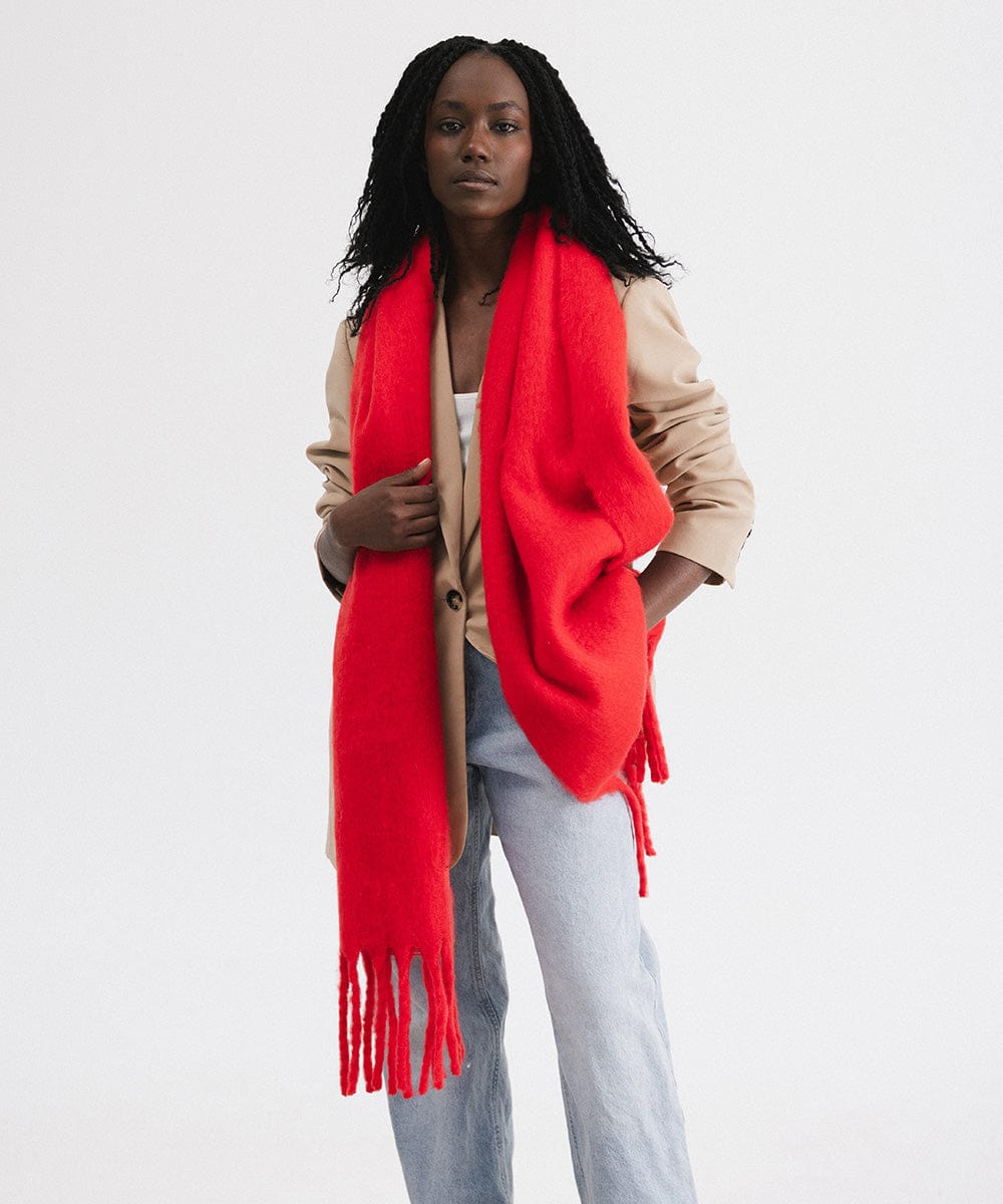 A person wearing a beige blazer, blue jeans, and a red oversized knit scarf with fringes, draped over the neck against a plain background #color_red