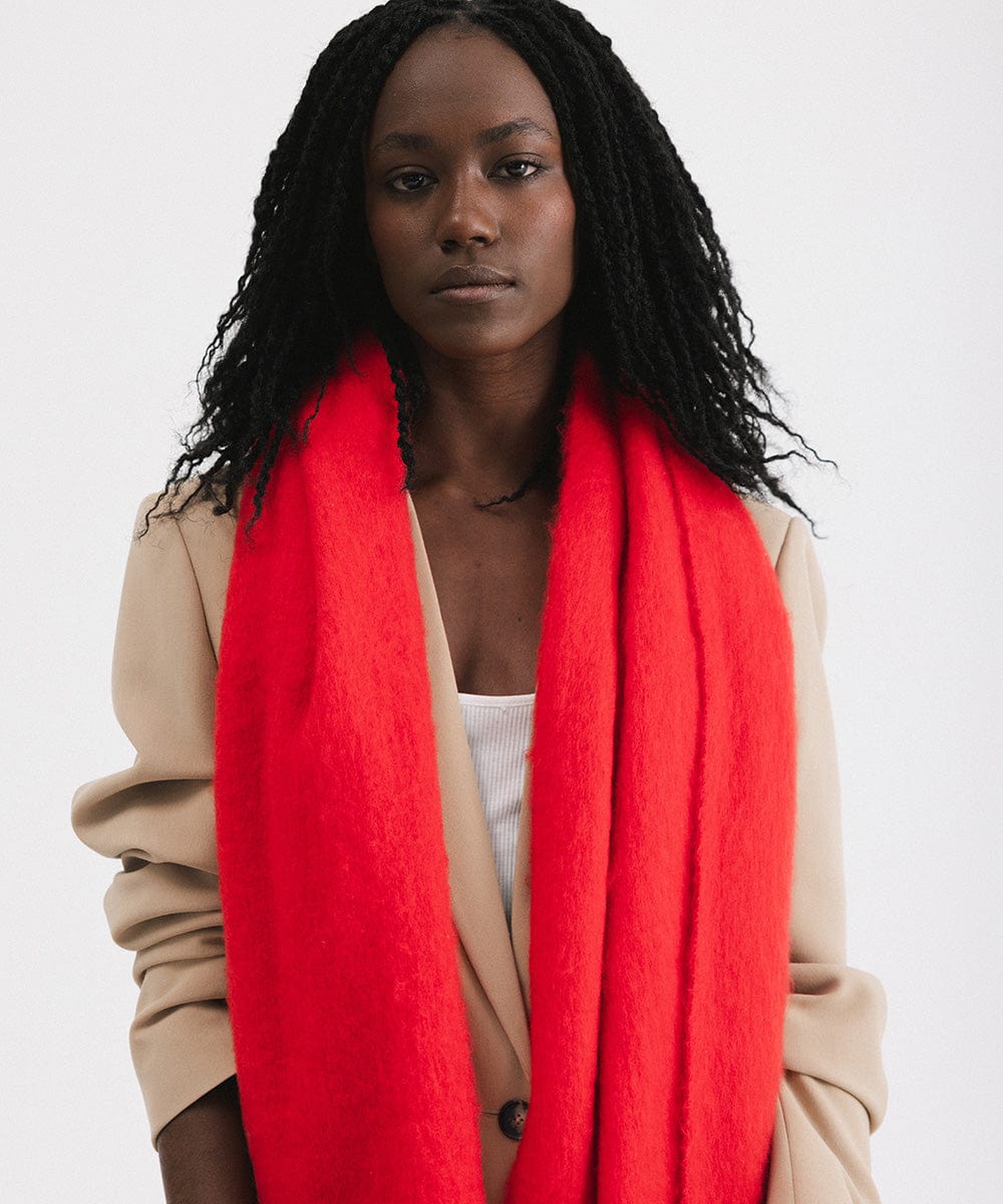 A person wearing a beige blazer, and a red oversized knit scarf with fringes, draped over the neck against a plain background #color_red