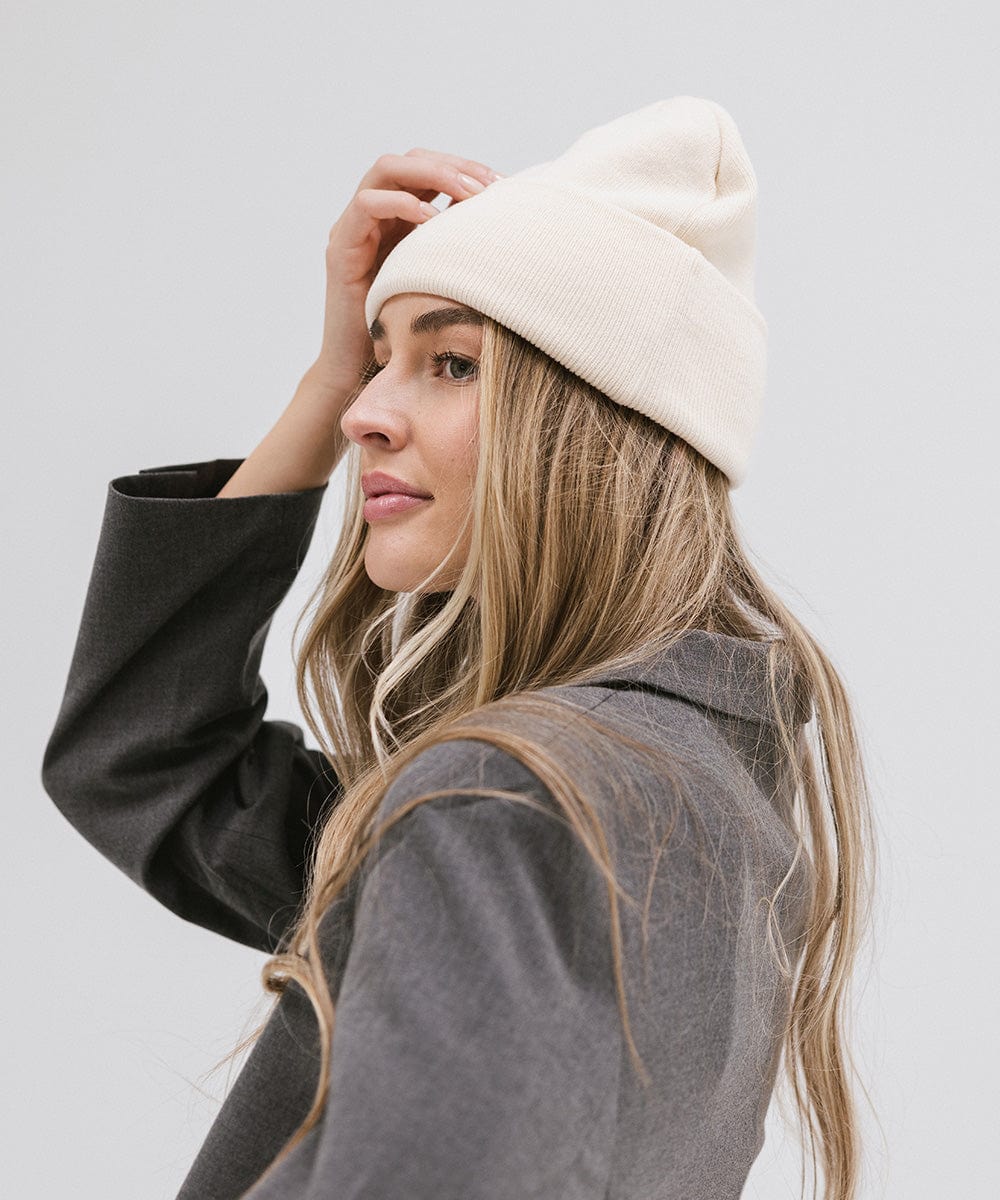 Woman wearing a cream beanie and a grey coat, posing sideways against a plain background #color_cream