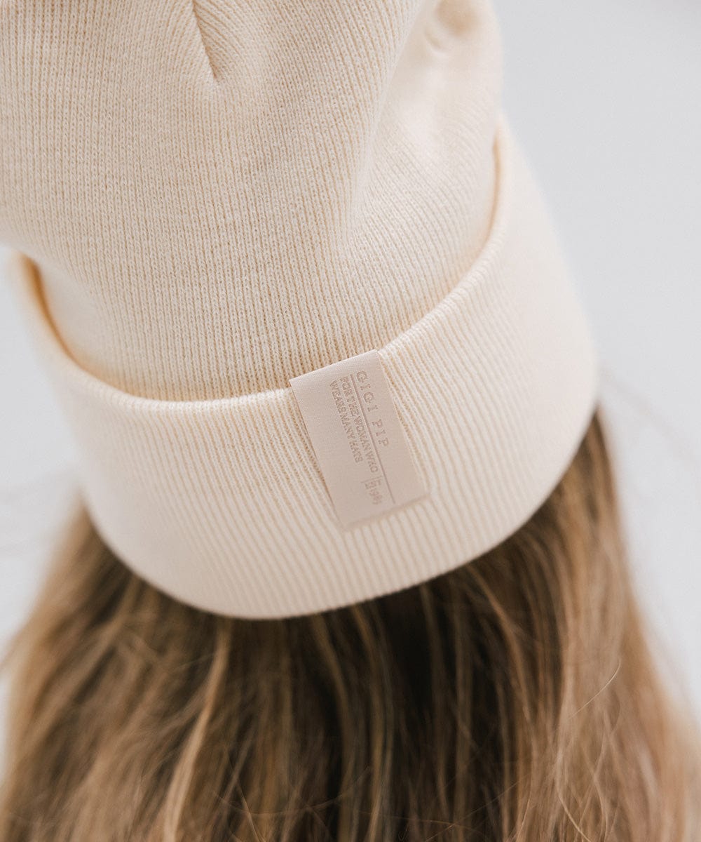 Close up image of someone wearing a cream beanie with the brand label against a plain background #color_cream
