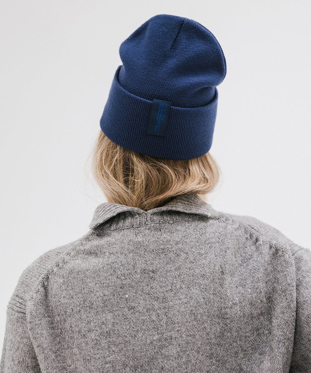 Woman wearing a navy beanie and a grey sweater, facing behind against a plain background #color_navy