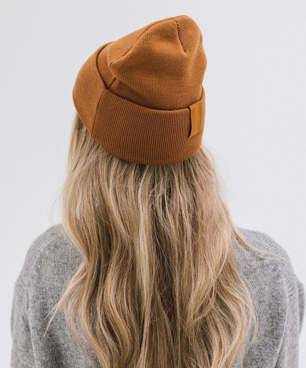 Woman with long hair wearing a cocoa colored beanie and a grey sweater, facing behind against a plain background #color_cocoa