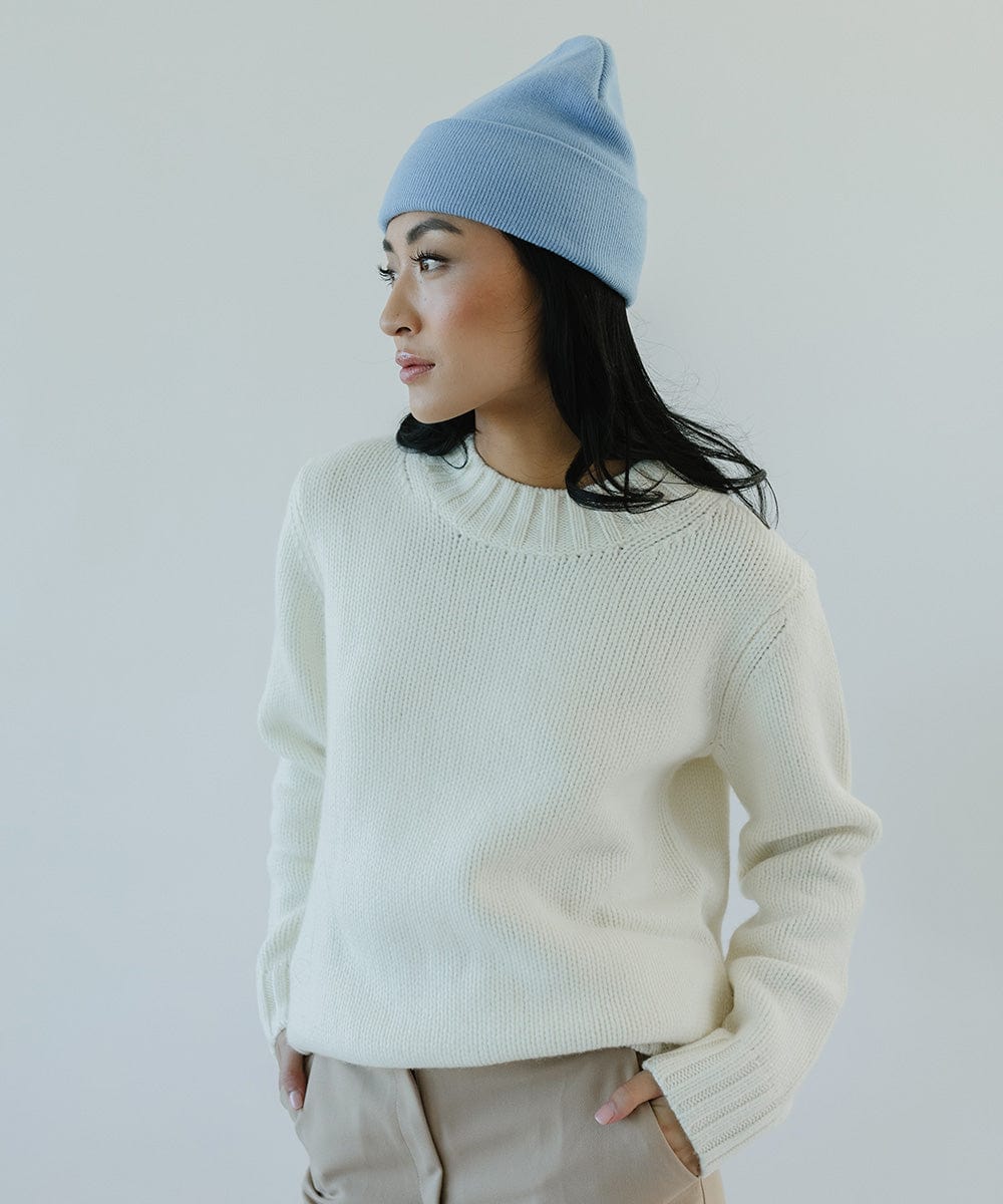 Woman wearing an azure blue colored beanie, a white sweater and beige pants, facing sideways against a plain background #color_azure blue
