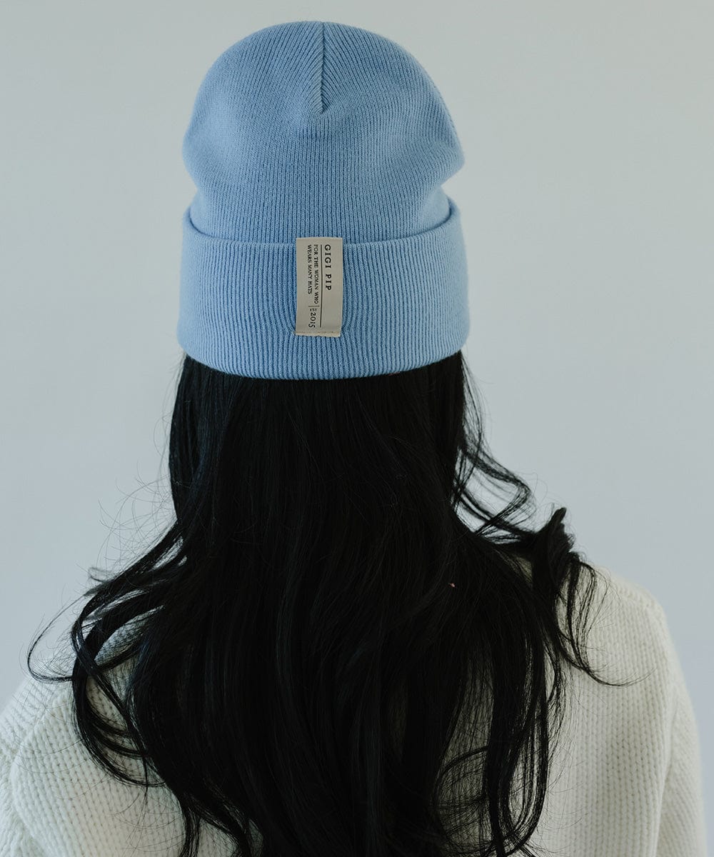 Woman with long hair wearing an azure blue colored beanie with the brand label and a white sweater, facing behind against a plain background #color_azure blue