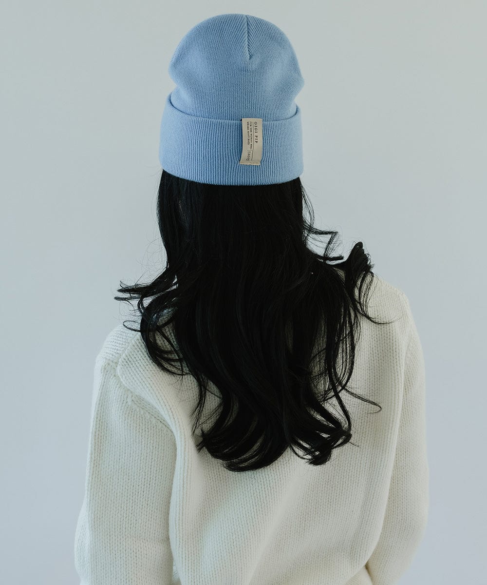 Woman with long hair wearing an azure blue colored beanie and a white sweater, facing behind against a plain background #color_azure blue