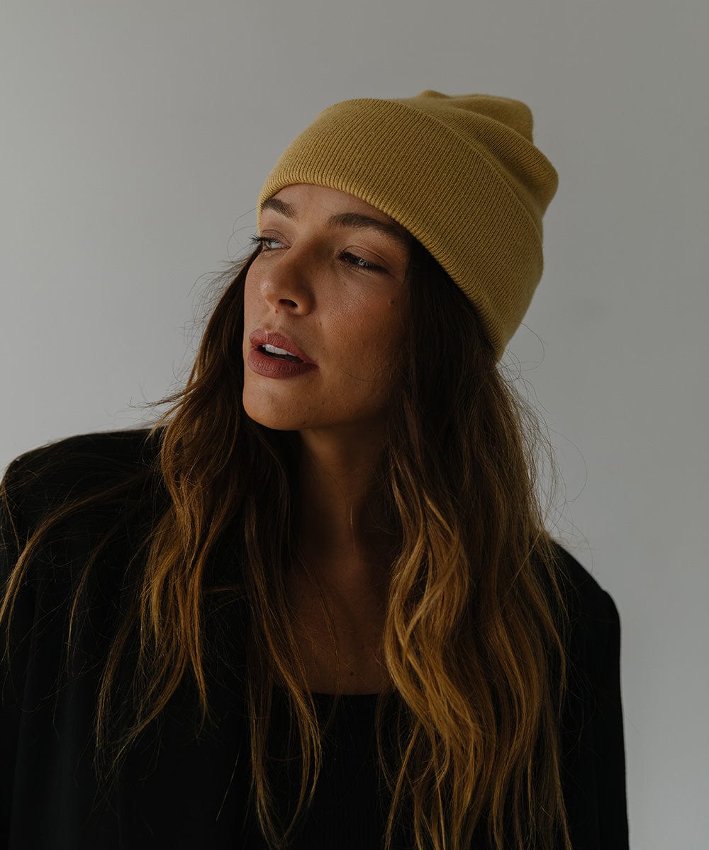Woman wearing a honeycomb colored beanie and a black coat, posing sideways against a plain background #color_honeycomb