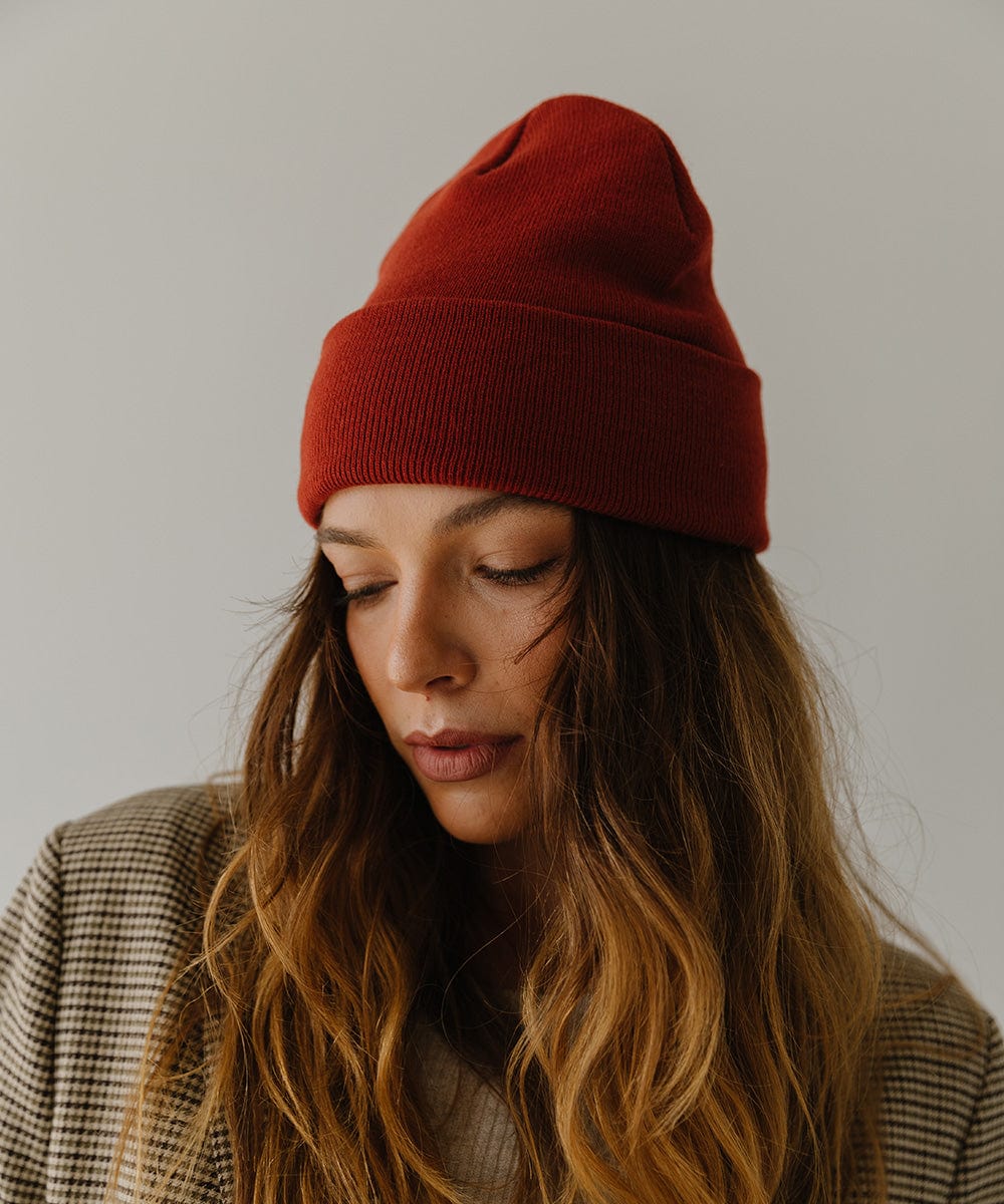 Woman wearing a brick red colored beanie and a coat, posing against a plain background #color_brick red