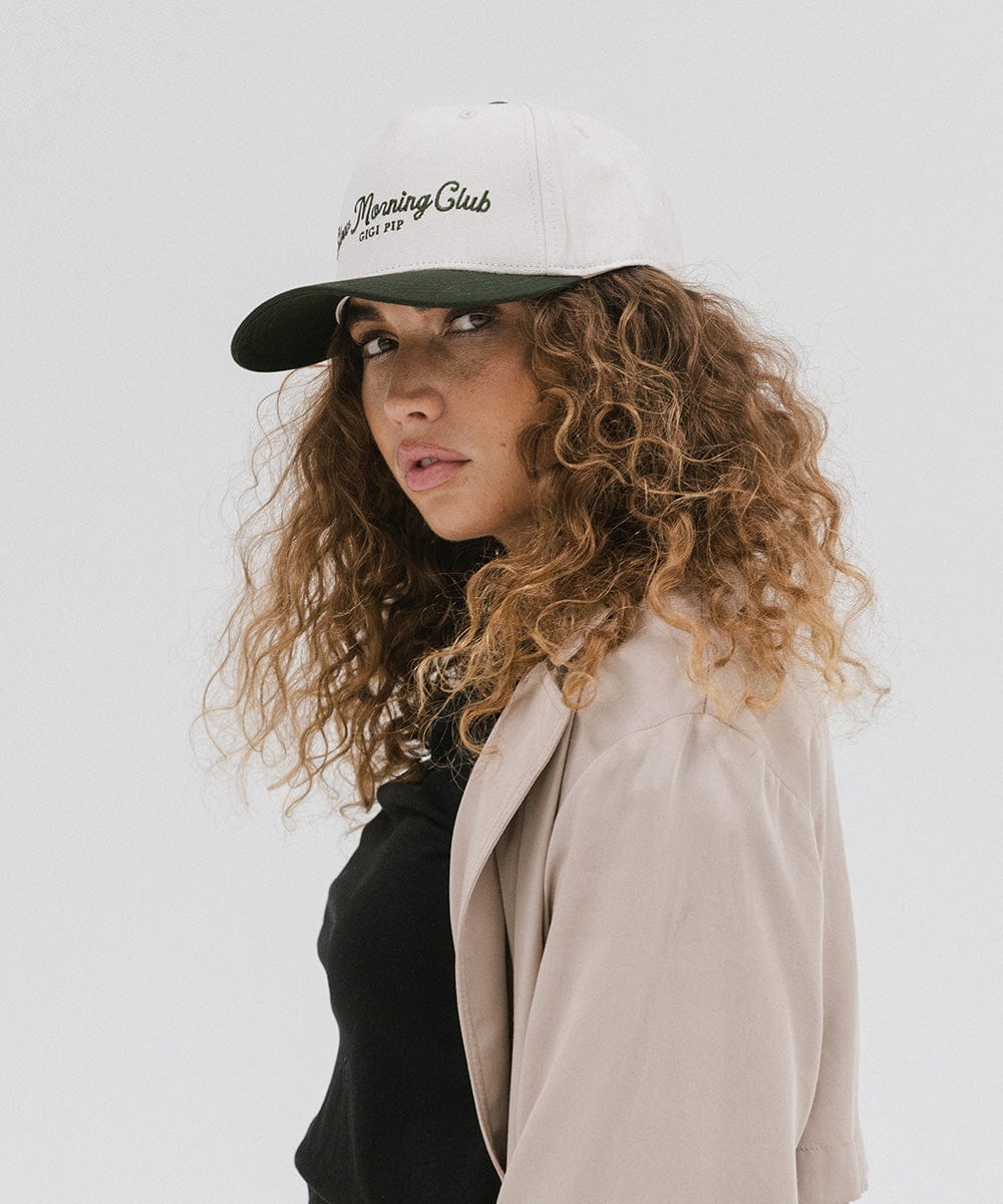 Woman wearing a cream and dark green cap with 'Slow Morning Club' text and a black top with a beige coat against a plain background #color_cream - dark green