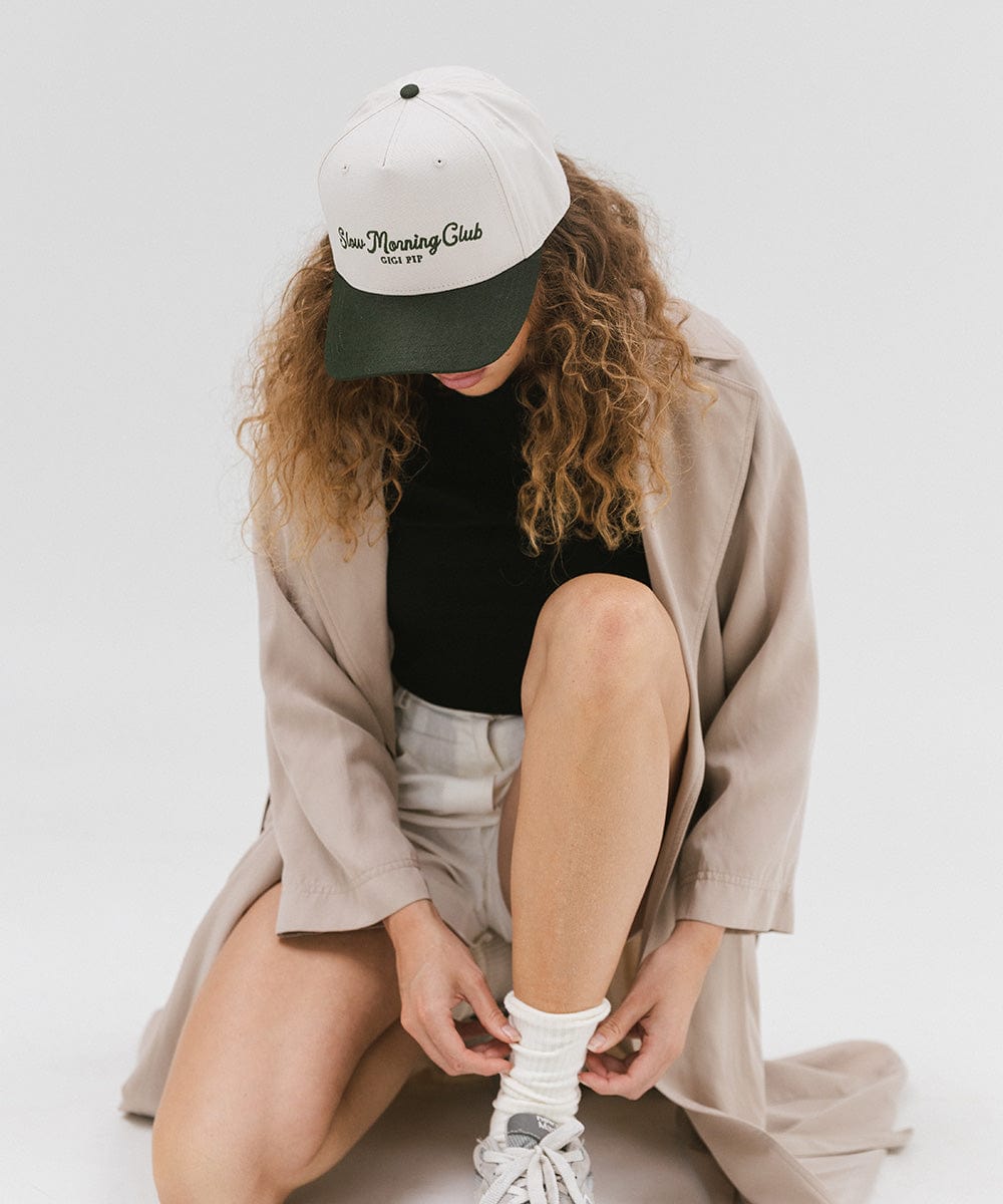 Woman wearing a cream and dark green cap with 'Slow Morning Club' text, a black top with a beige coat, and white shorts, sitting against a plain background #color_cream - dark green