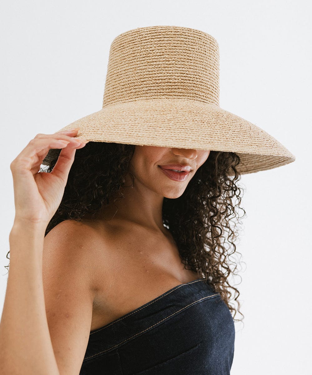 Woman with curly hair wearing a natural colored oversized packable straw bucket hat and a denim top, posing against a plain background #color_natural