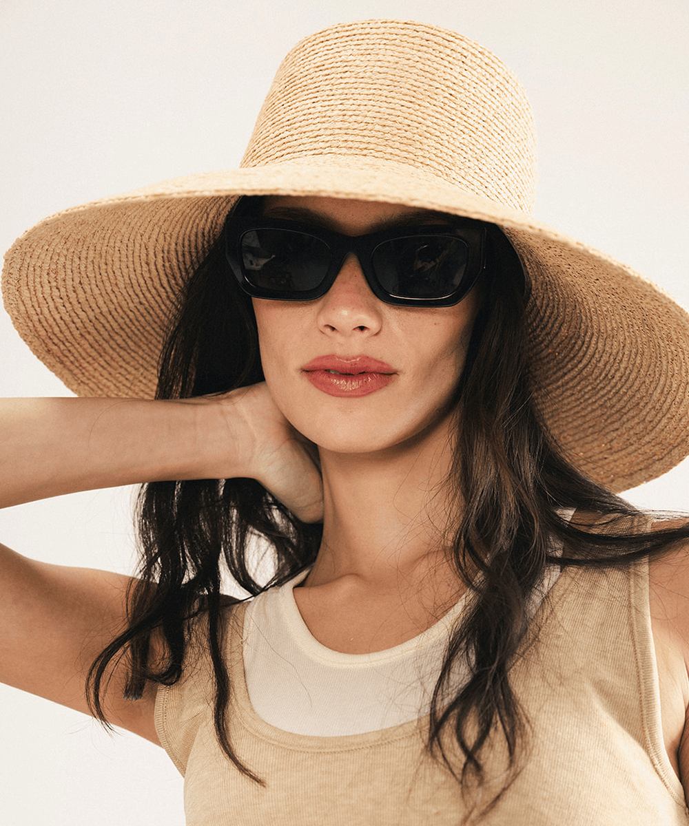 Woman wearing a natural colored oversized raffia straw bucket hat with a wide brim and sunglasses on a plain background #color_natural