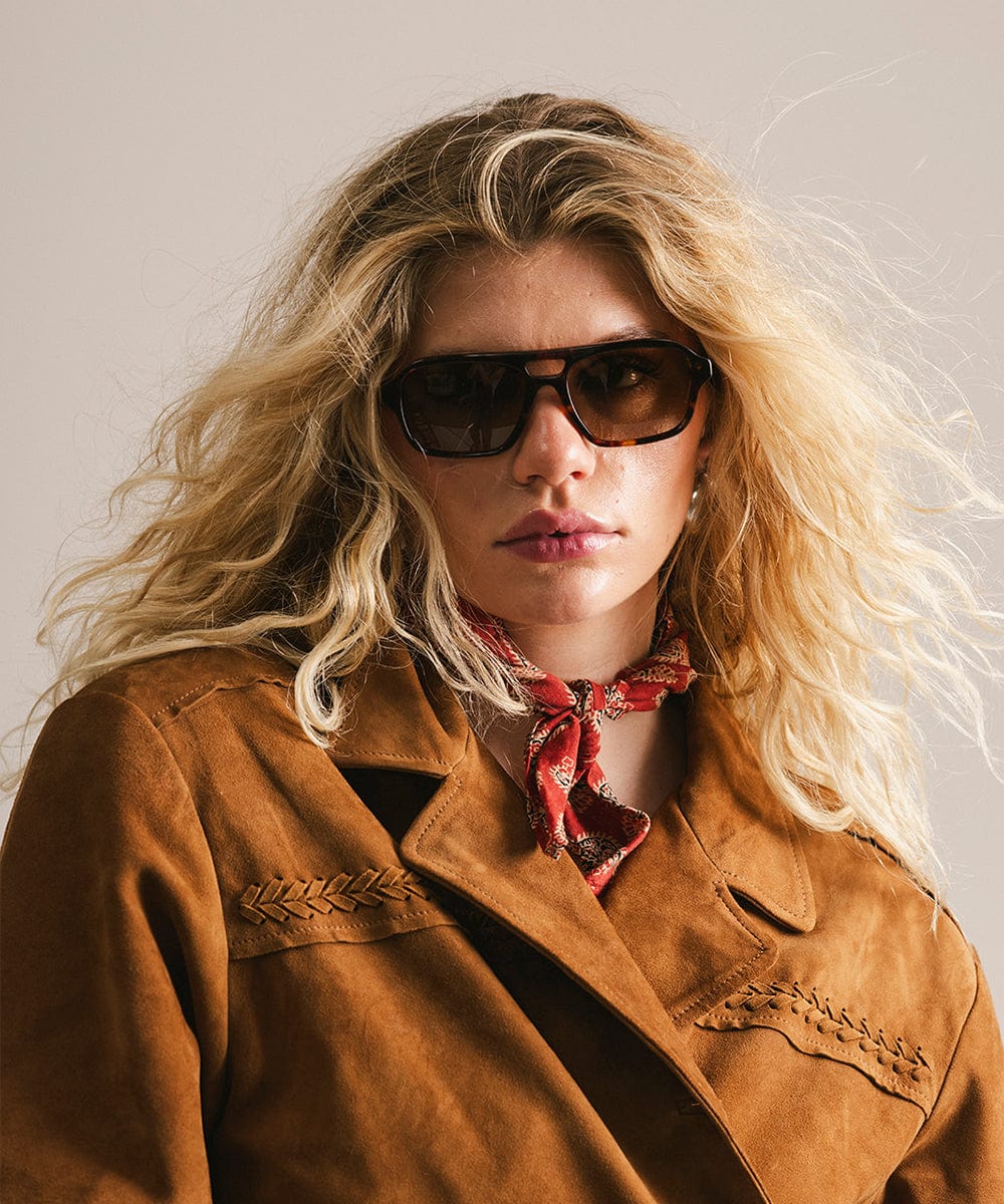Woman wearing sunglasses and a brown jacket with a red scarf, against a neutral background #color_tortoise