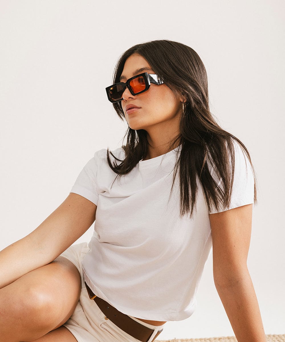 Woman wearing sunglasses with black frame and orange lenses and a white t-shirt with beige shorts, sitting against a plain background #color_black