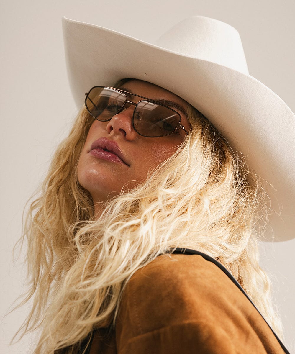 Person wearing a white cowboy hat and brown sunglasses, posing sideways against a neutral background #color_brown