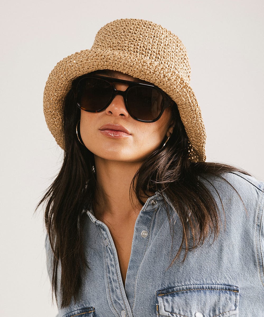 Woman wearing brown hat, tortoise colored aviator sunglasses and a denim shirt against a plain background #color_tortoise