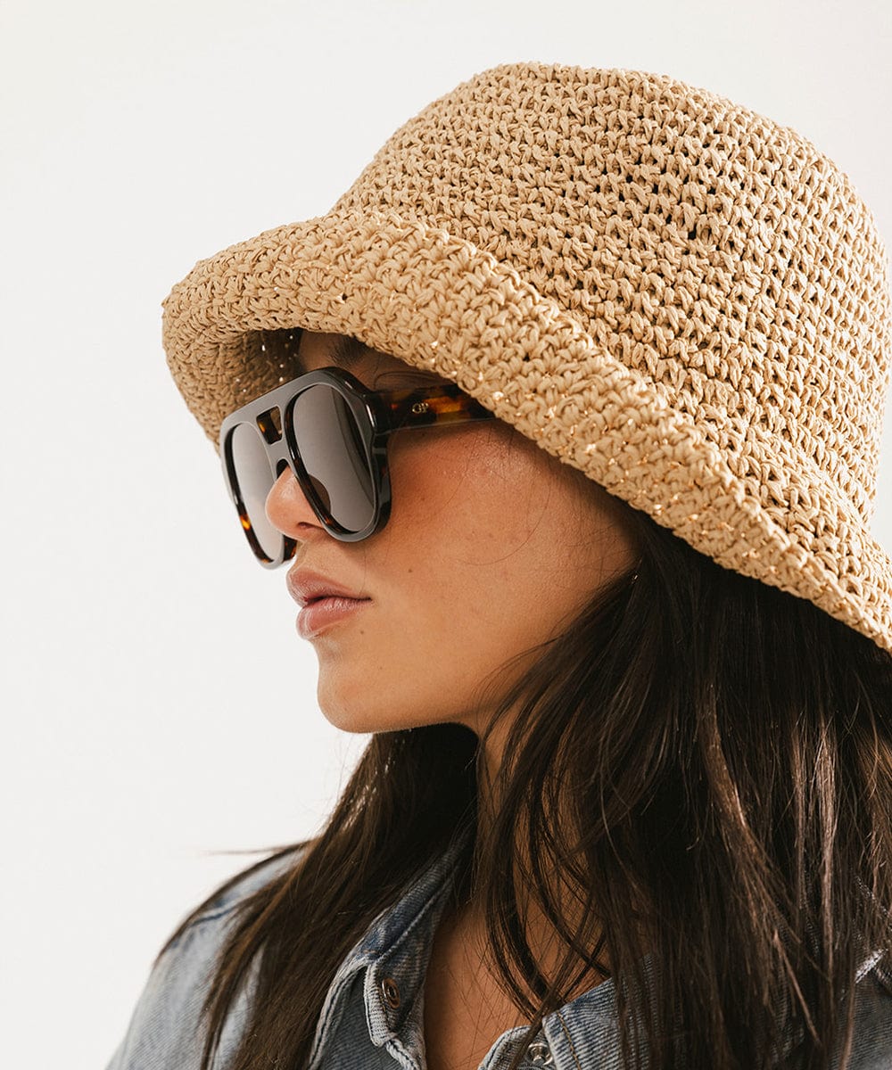 Woman wearing a brown hat and tortoise colored aviator sunglasses, facing sideways against a plain background #color_tortoise