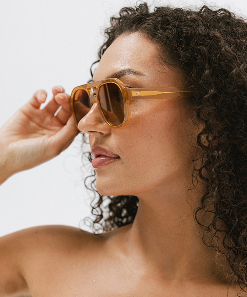 Woman with curly hair wearing honey colored aviator sunglasses against a plain background #color_honey