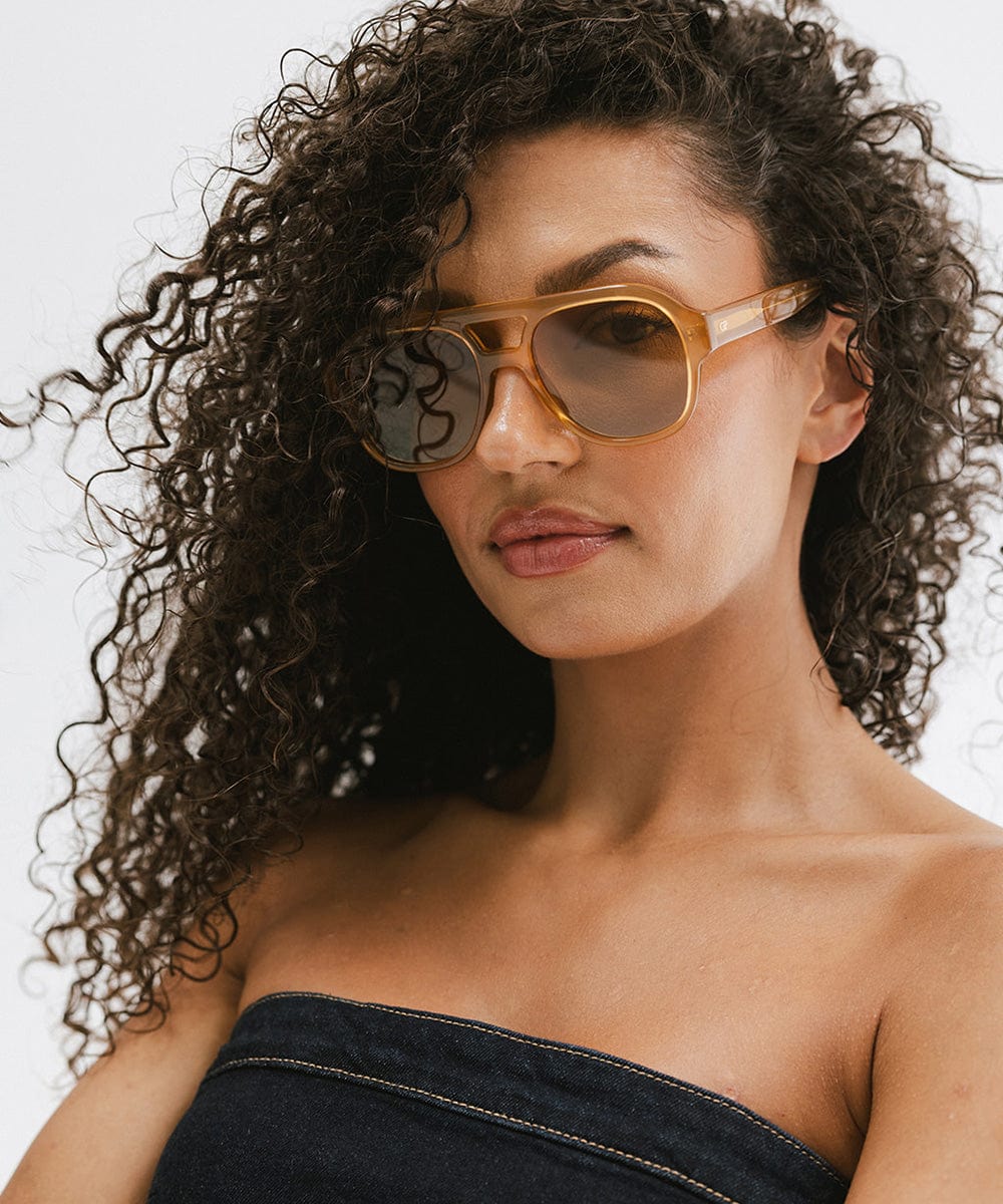 Woman with curly hair wearing honey colored aviator sunglasses and a denim top against a plain background #color_honey