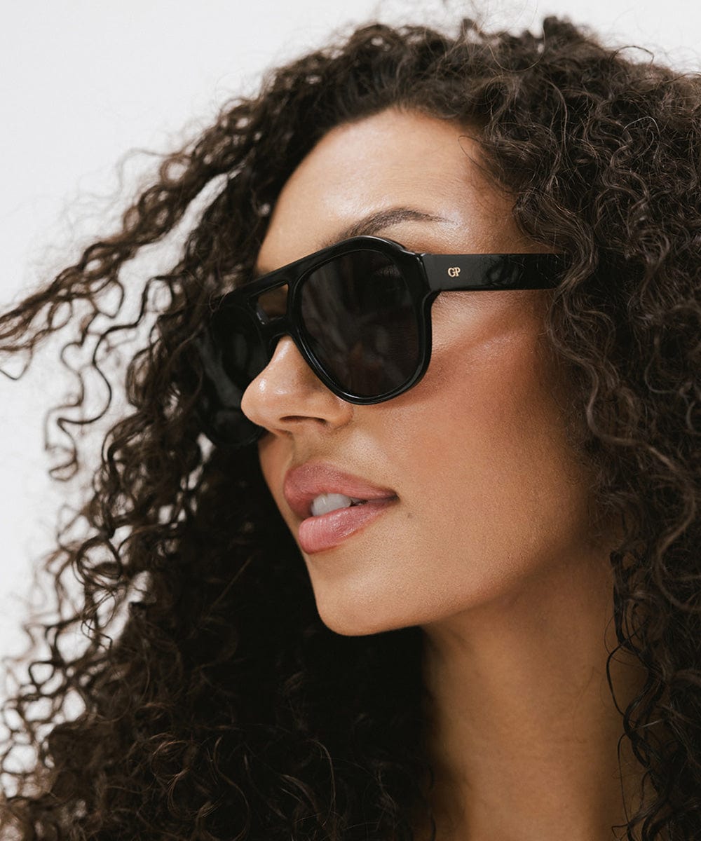Woman with curly hair wearing black aviator sunglasses against a plain background #color_black