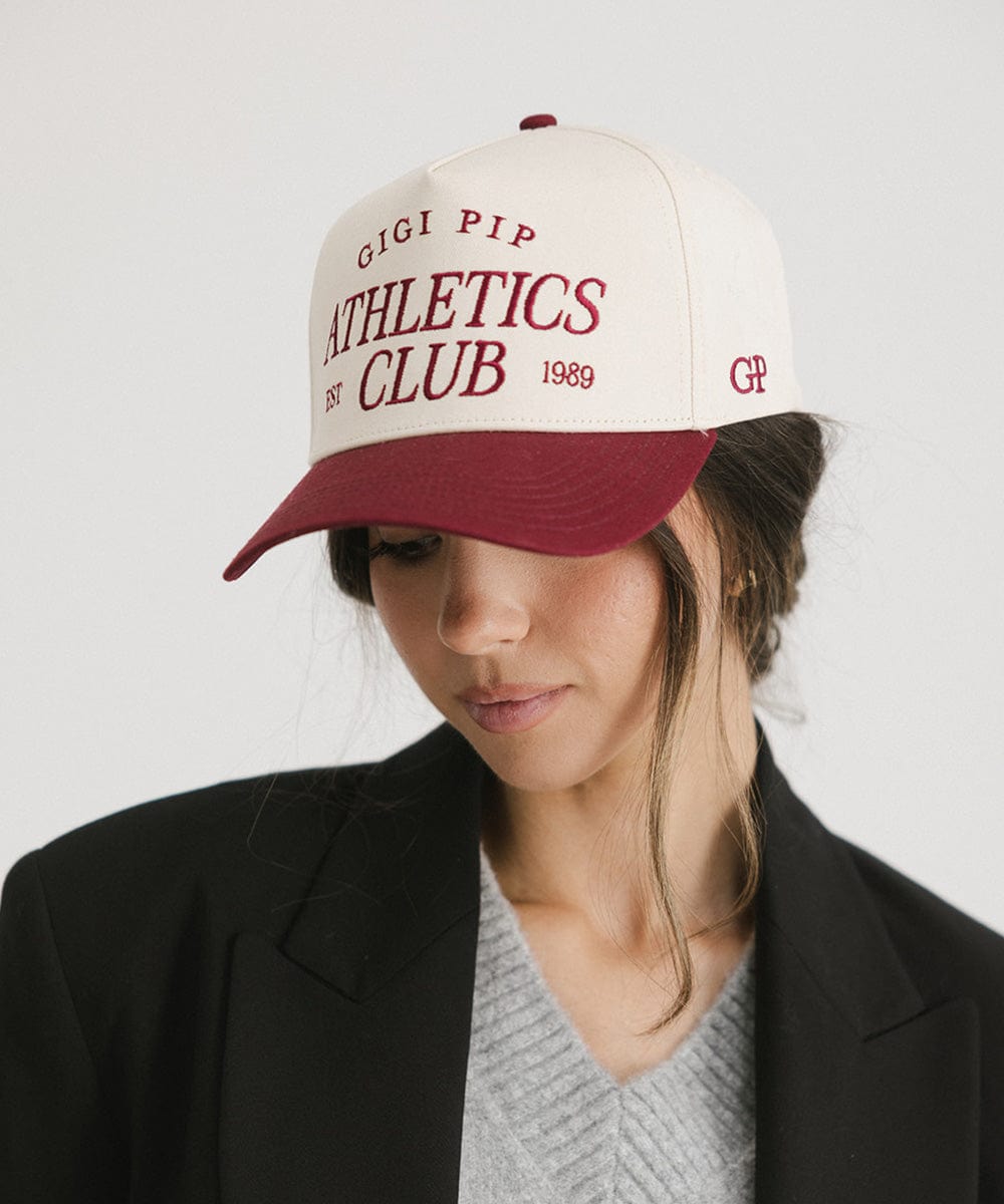 Woman wearing a Burgundy Trucker hat with 'Gigi Pip Athletics Club' text and a black coat against a plain background #color_burgundy