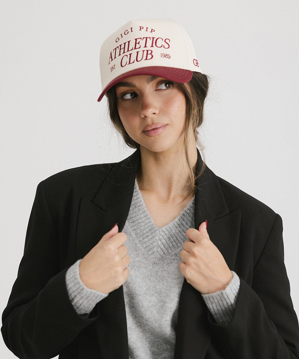 Woman wearing a Burgundy Trucker hat with 'Gigi Pip Athletics Club' text and a grey sweater with a black coat against a plain background #color_burgundy