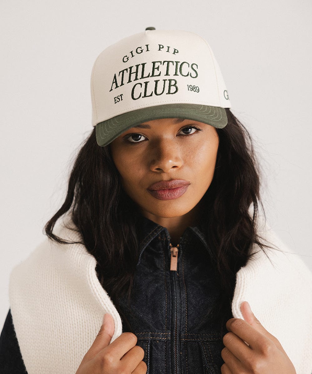 Woman wearing a cream and dark green Trucker hat with 'Gigi Pip Athletics Club' text and a denim jacket against a plain background #color_cream - dark green