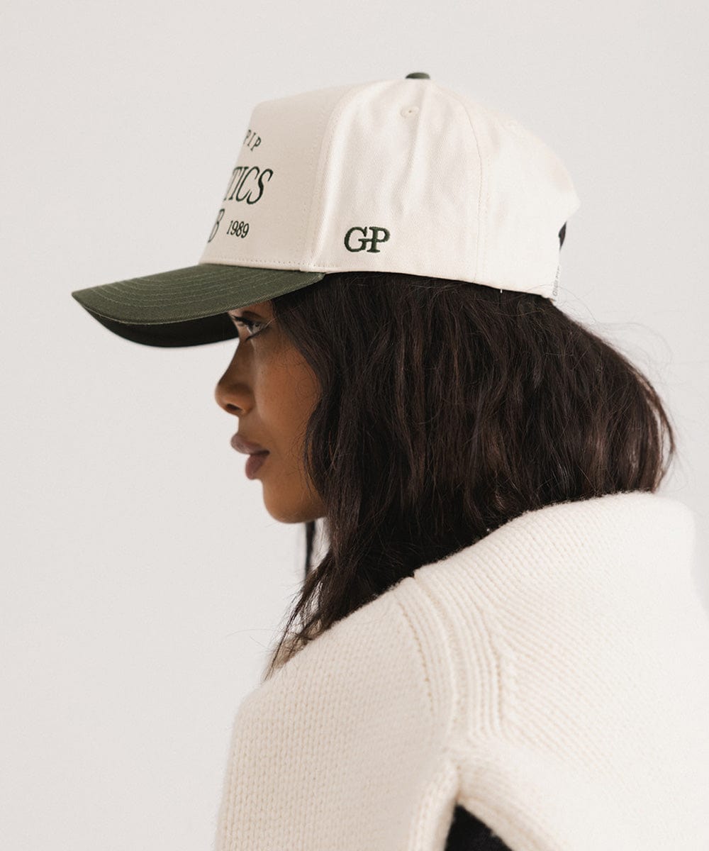 Woman wearing a cream and dark green Trucker hat and a white sweater, facing sideways against a plain background #color_cream - dark green