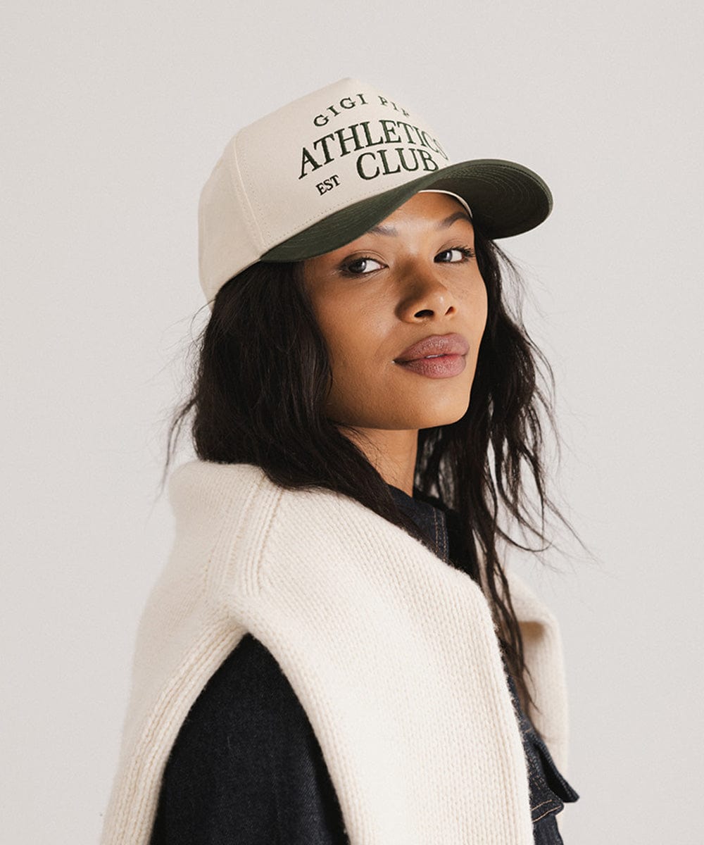 Woman wearing a cream and dark green Trucker hat with 'Gigi Pip Athletics Club' text and a denim jacket, posing sideways against a plain background #color_cream - dark green